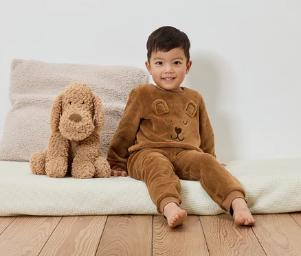 Un garçon souriant en pyjama marron à motif d'ours est assis à côté d'un chien en peluche sur un coussin blanc.