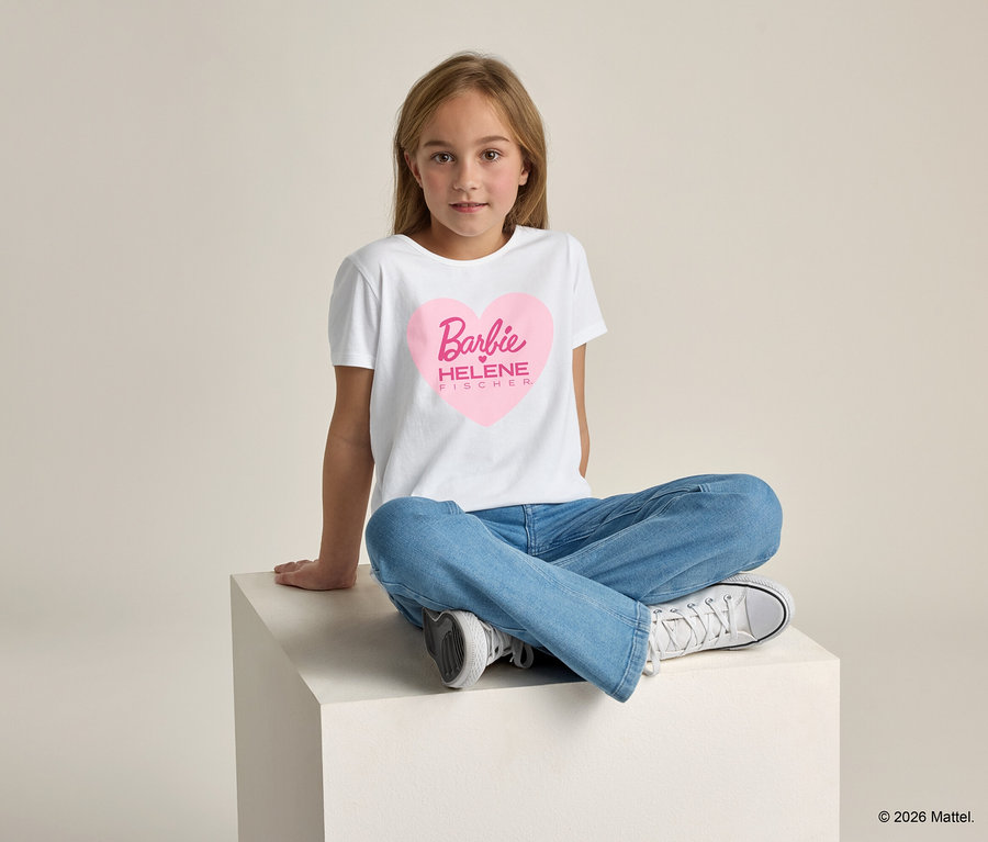 Une fille est assise les jambes croisées sur un cube blanc et porte un t-shirt blanc avec un cœur rose sur lequel est écrit « Barbie HELENE FISCHER ».