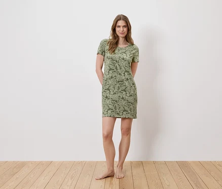 Une femme se tient pieds nus devant un mur blanc, portant une robe verte à fleurs à manches courtes.