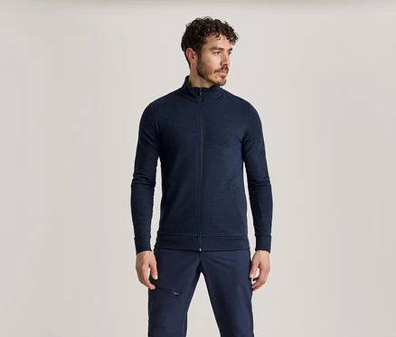 Un homme aux cheveux bouclés foncés, avec une barbe et une moustache, porte une veste zippée bleu marine et un pantalon bleu marine.