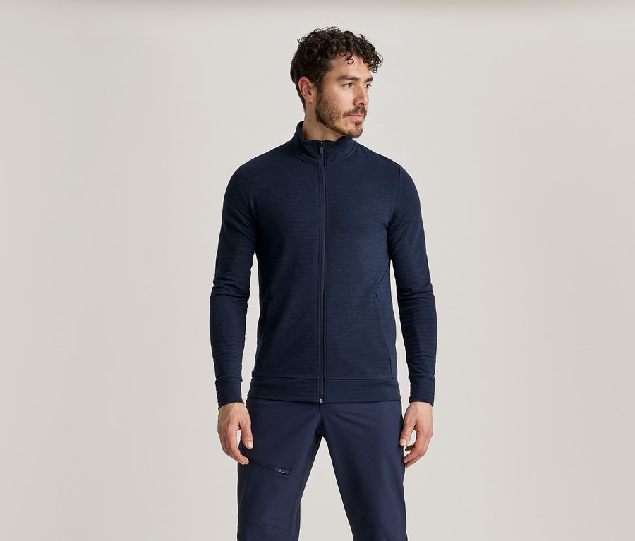 Un homme aux cheveux bouclés foncés, avec une barbe et une moustache, porte une veste zippée bleu marine et un pantalon bleu marine.