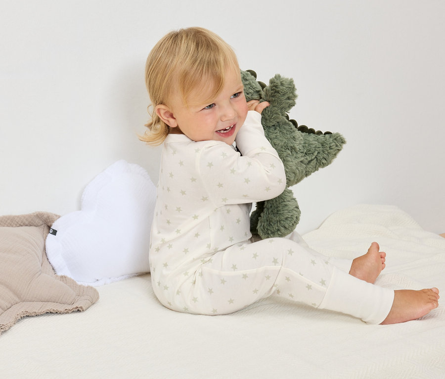Un enfant blond en pyjama blanc à étoiles est assis sur une couverture blanche et serre dans ses bras une peluche de dinosaure verte.