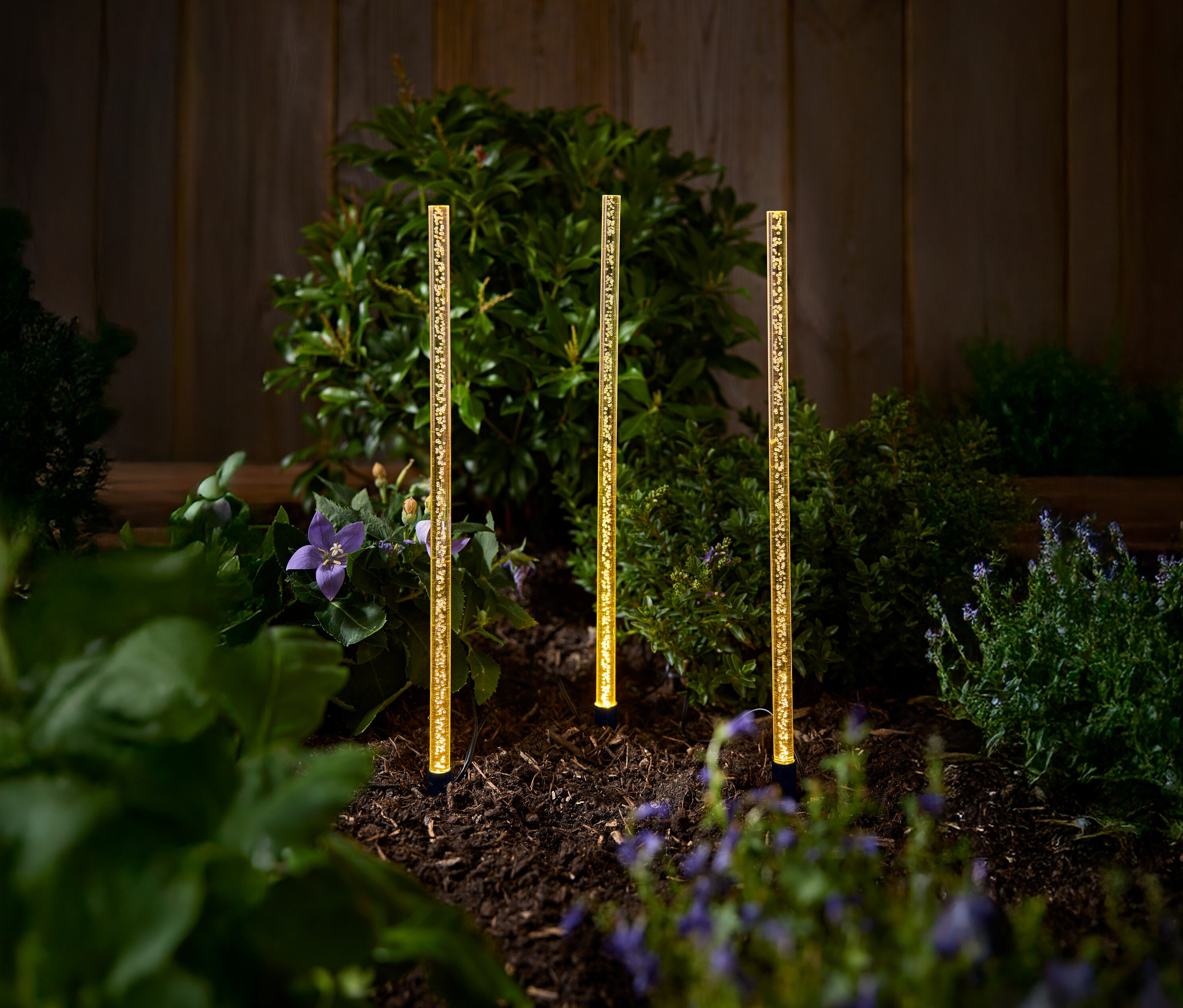 Trois lampes solaires de jardin dorées sont posées sur un parterre de fleurs avec des buissons et des fleurs.