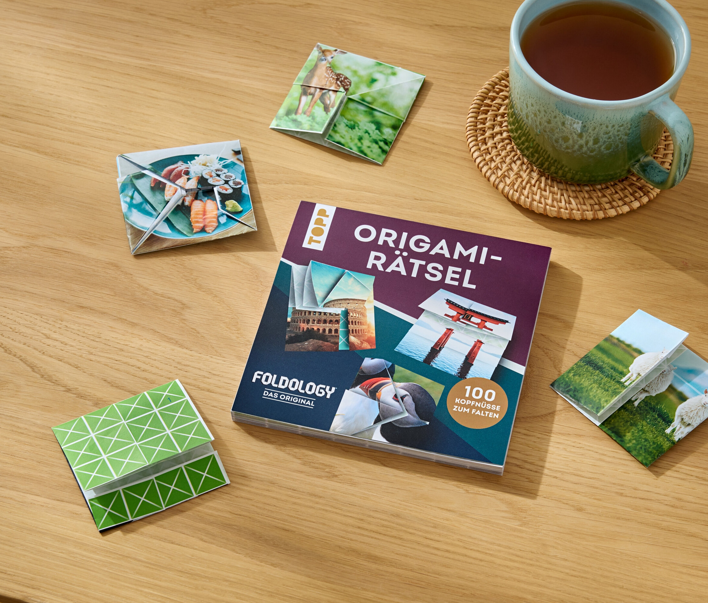 Sur une table en bois sont posés le livre »Origami-Rätsel«, une tasse de thé et des figurines origami pliées avec différents motifs.
