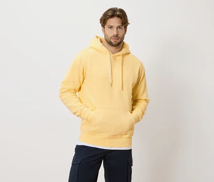Un homme aux cheveux châtain clair et à la barbe porte un sweat à capuche jaune et un pantalon cargo bleu marine sur un fond blanc.