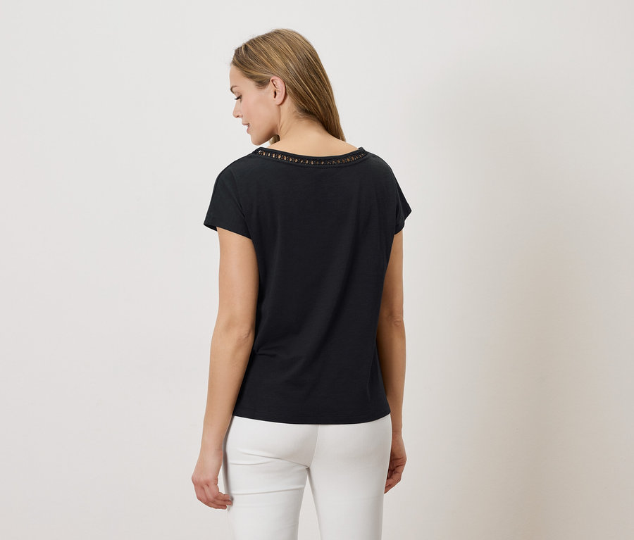 Femme vue de dos, portant un t-shirt noir avec un motif ajouré au niveau de l'encolure et un pantalon blanc sur fond blanc.