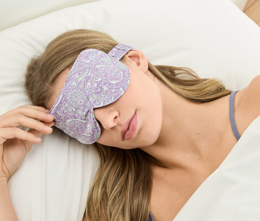 Femme allongée sur un oreiller blanc avec un masque de sommeil violet à motifs.
