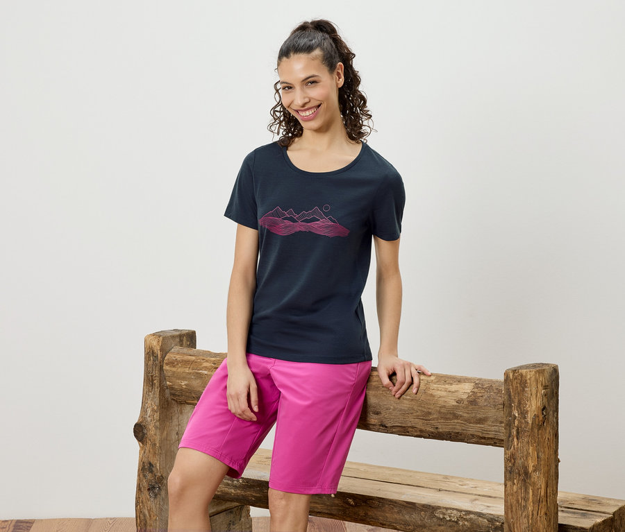 Femme appuyée sur un banc en bois, portant un t-shirt bleu marine avec un motif de montagne rose et un short rose.
