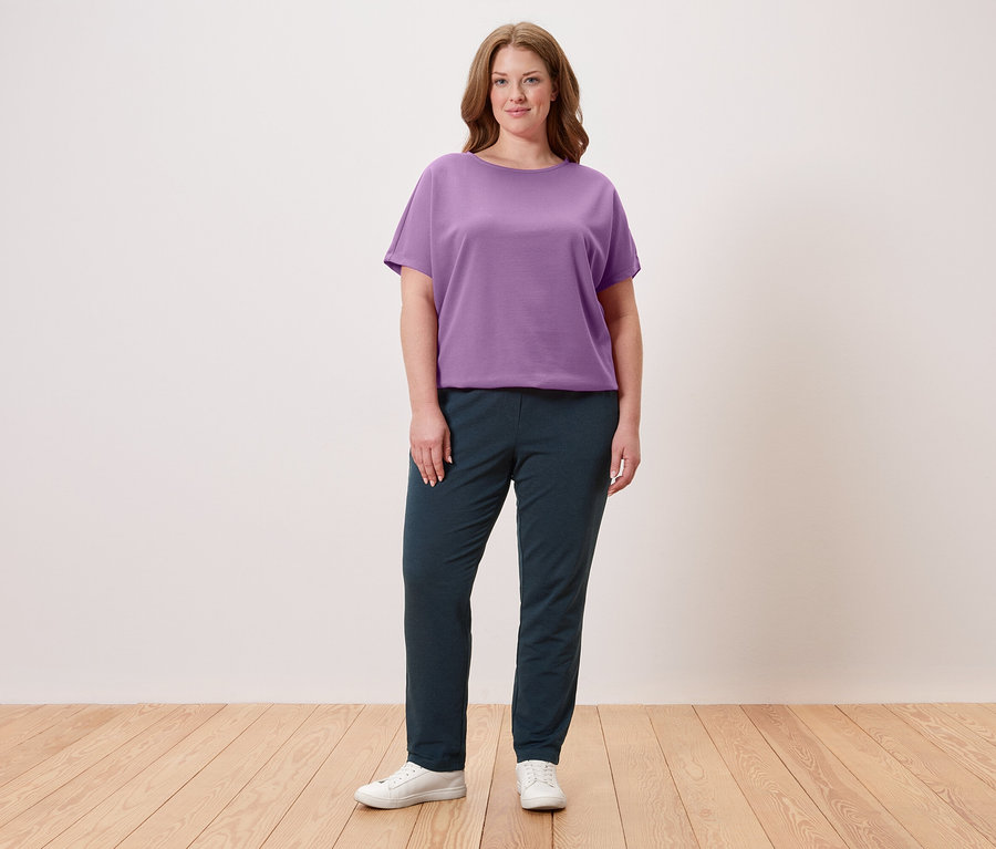 Une femme se tient devant un mur, portant un t-shirt violet, un pantalon foncé et des baskets blanches.