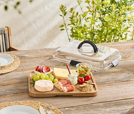 Sur une table en bois, une planche en bois avec du fromage, de la saucisse, des olives et des crackers, partiellement recouverte d'un couvercle transparent.
