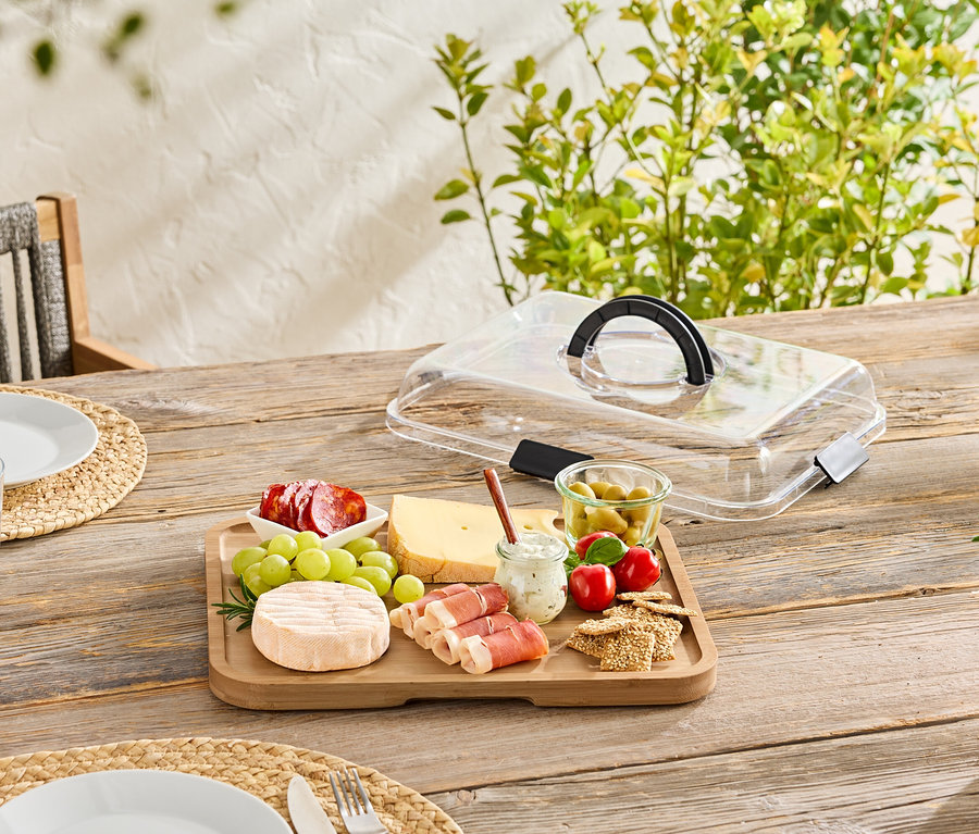 Sur une table en bois, une planche en bois avec du fromage, de la saucisse, des olives et des crackers, partiellement recouverte d'un couvercle transparent.