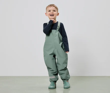 Un garçon souriant en salopette verte et bottes en caoutchouc se tient devant un fond clair.
