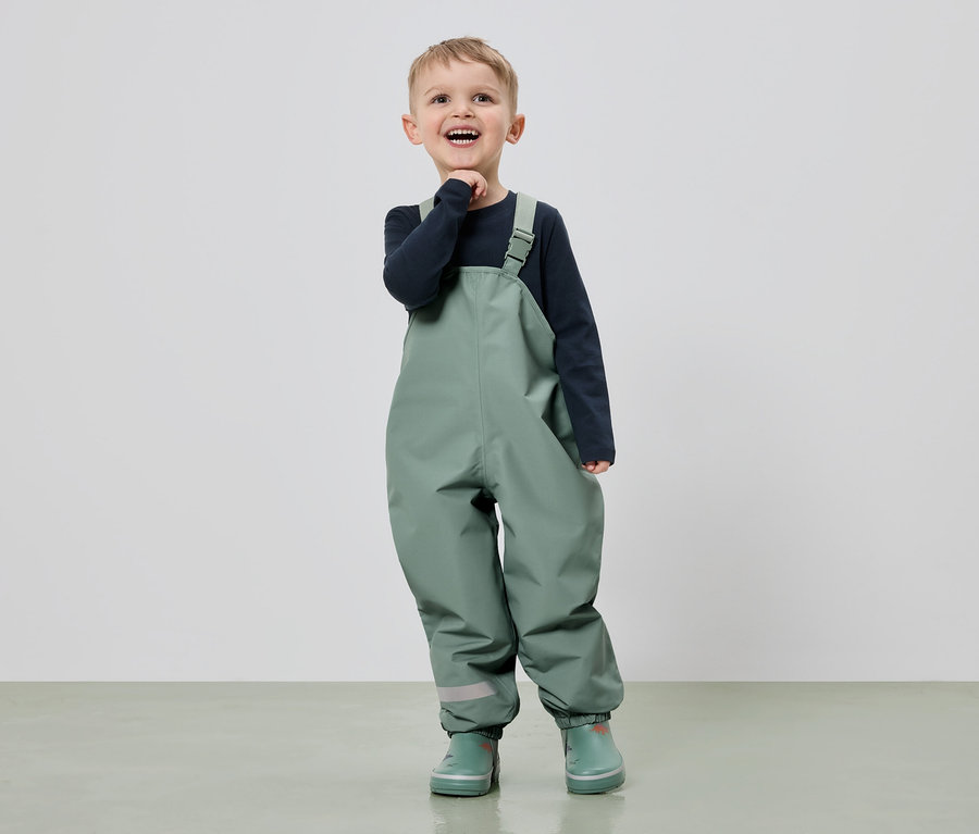 Un garçon souriant en salopette verte et bottes en caoutchouc se tient devant un fond clair.
