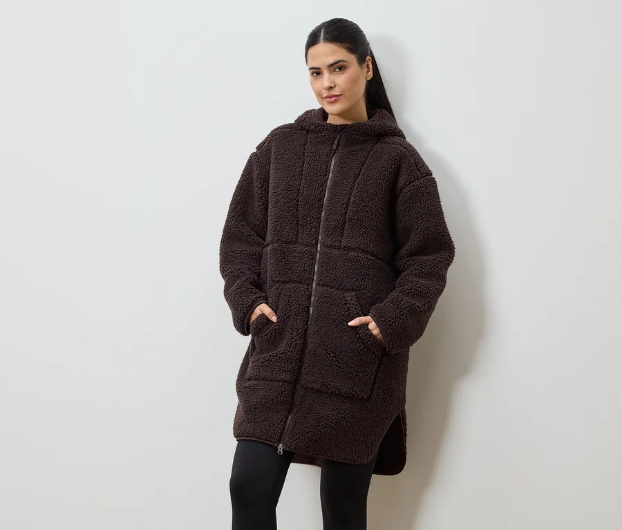 Une femme, les mains dans les poches, porte un long manteau en fausse fourrure marron à capuche et fermeture éclair sur un legging noir.
