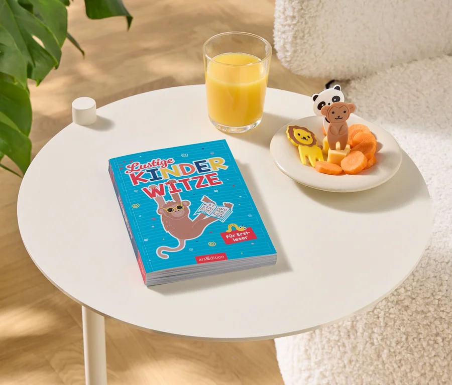 Sur une table se trouvent un livre intitulé "Lustige Kinderwitze", un verre de jus d'orange et une assiette avec des carottes et des figurines d'animaux.