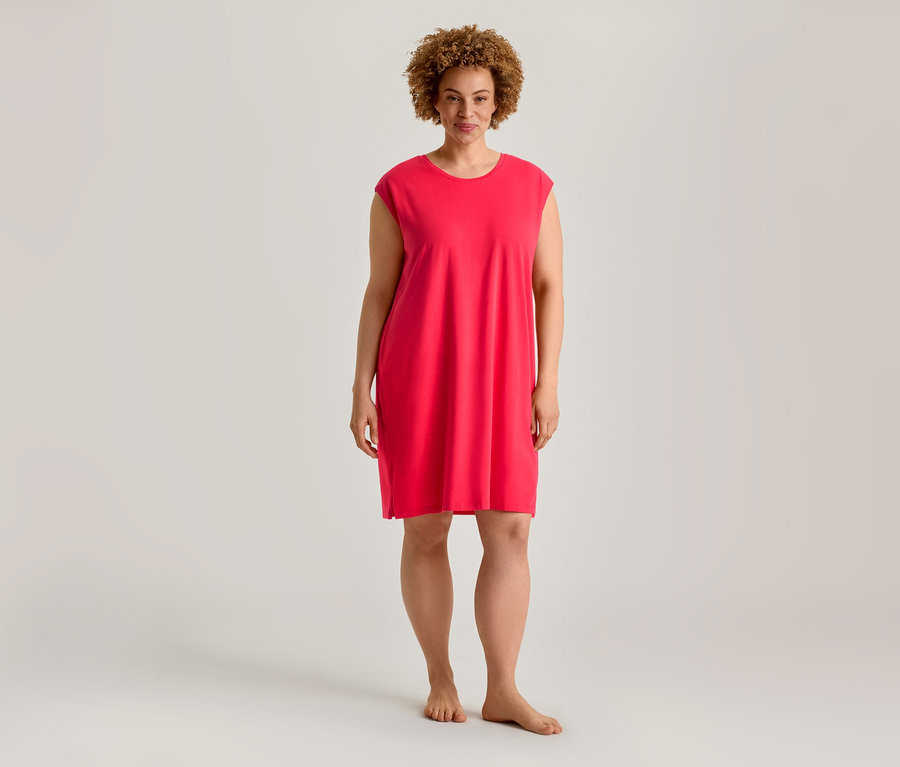 Une femme aux cheveux bouclés se tient pieds nus, vêtue d'une robe courte, ample et rose vif.