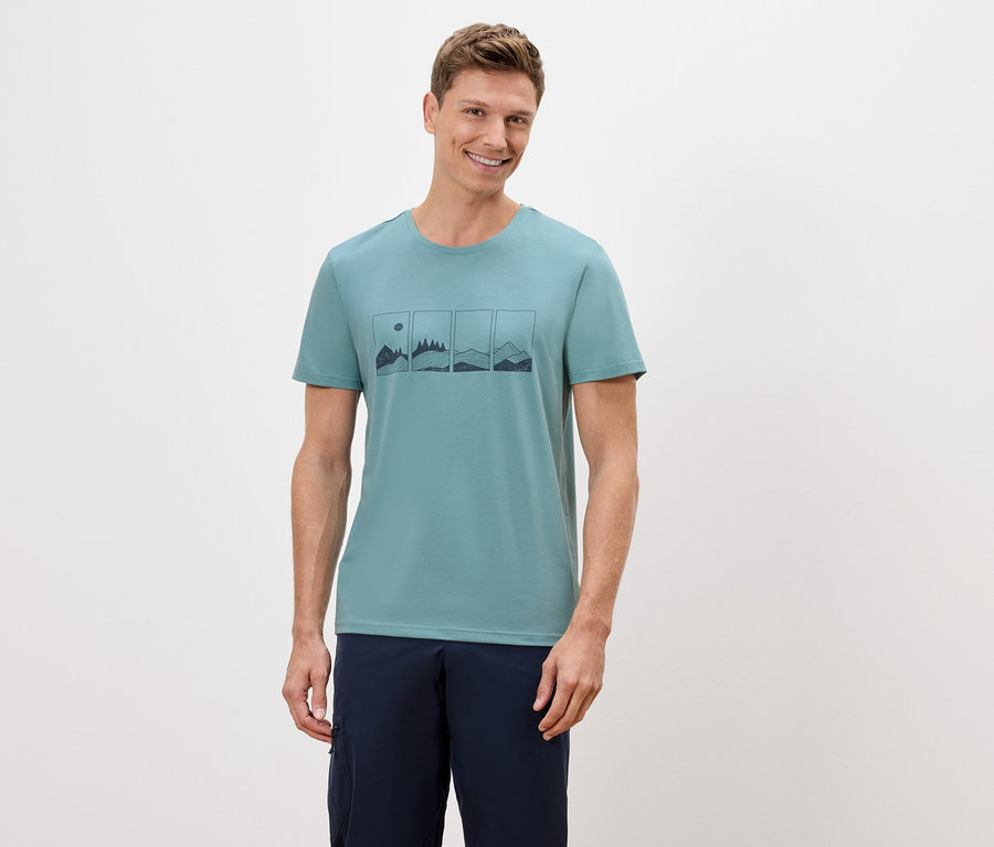 Un homme porte un t-shirt vert avec un motif de montagnes et de soleil.