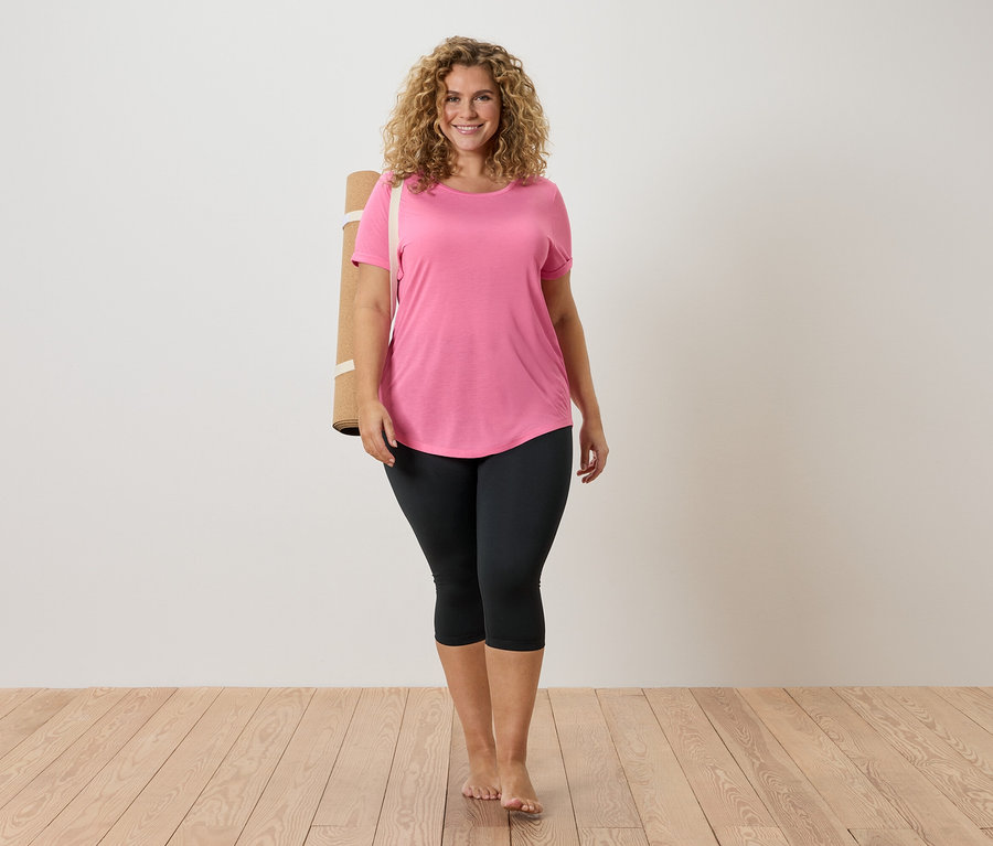 Femme aux cheveux bouclés portant un t-shirt rose et un legging noir, avec un tapis de yoga sur l'épaule, pieds nus sur un plancher en bois.