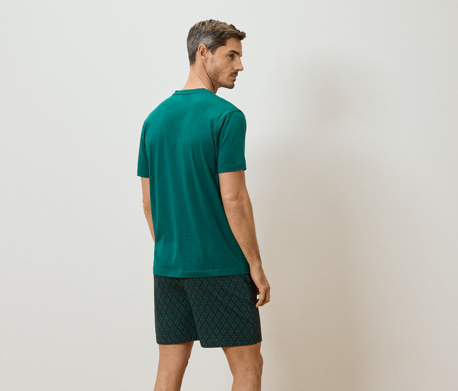 Un homme se tient dos à la caméra, portant un t-shirt vert et un short court à motifs.