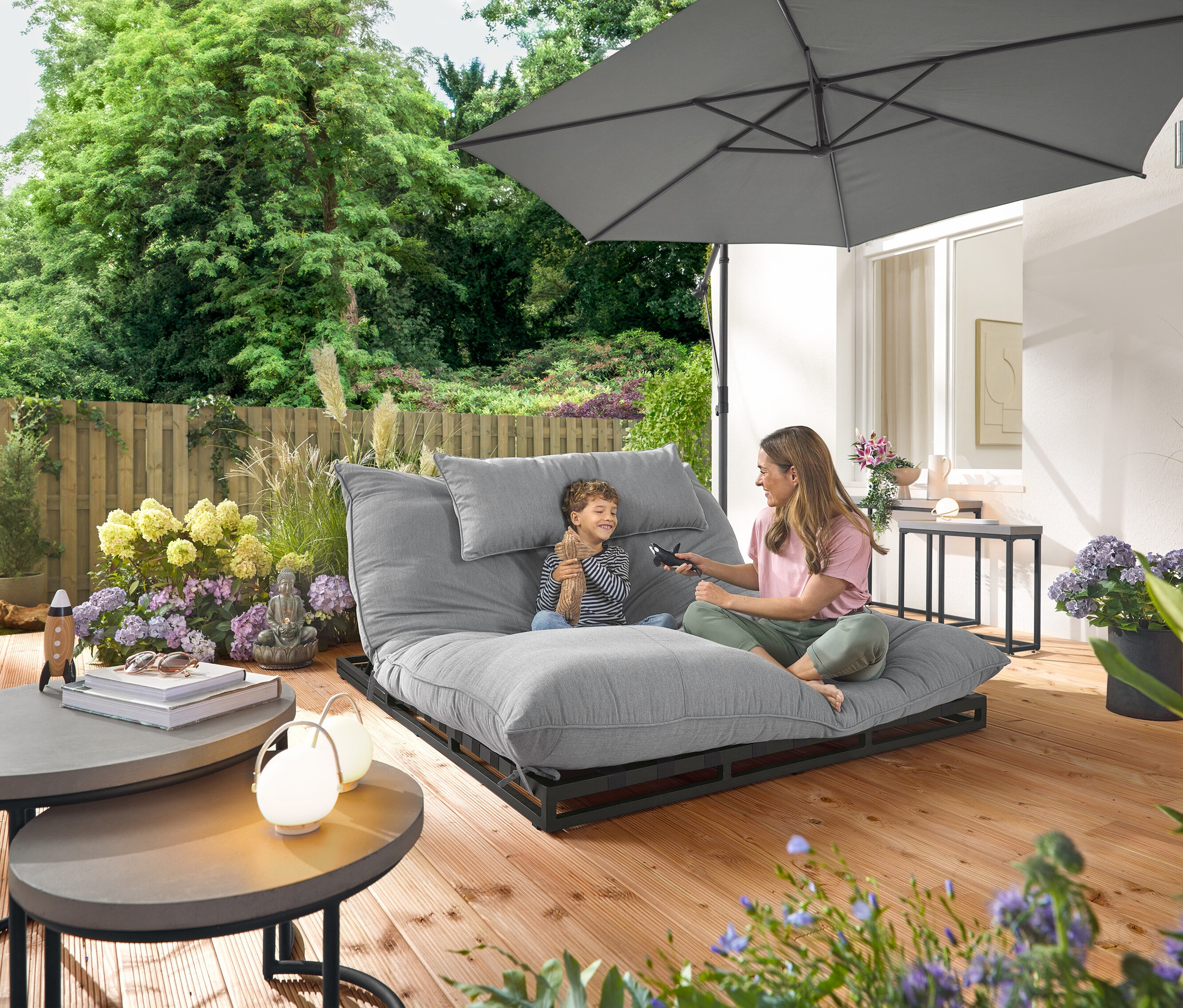 Une femme et un enfant sont assis sur une chaise longue double Premium-Doppel-Gartenliege avec tissu Creacryl®. Dans le jardin, il y a 2 tables basses Garten-Couchtische et 2 tables console Garten-Konsolentische avec plateaux en polyciment et 3 lampes de table Akku-Tischleuchten.