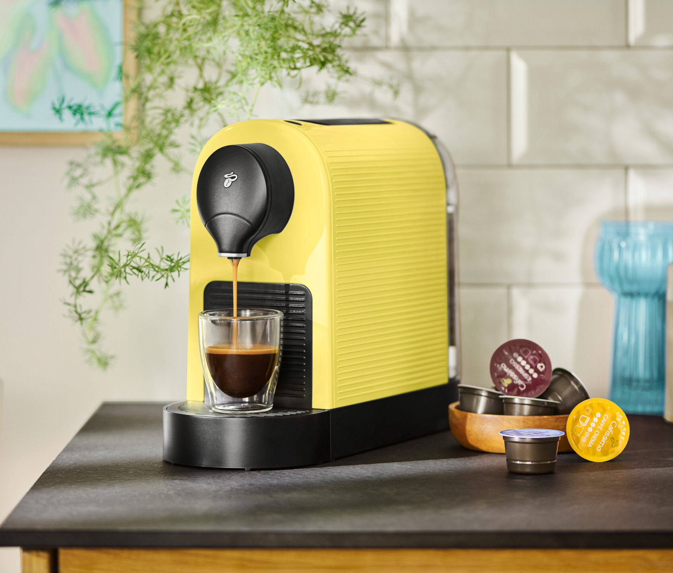 Une machine à café jaune remplit une tasse de café, des capsules sont à côté.