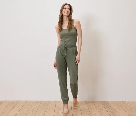 Une femme se tient pieds nus sur un plancher en bois, portant un débardeur et un pantalon de jogging vert olive.