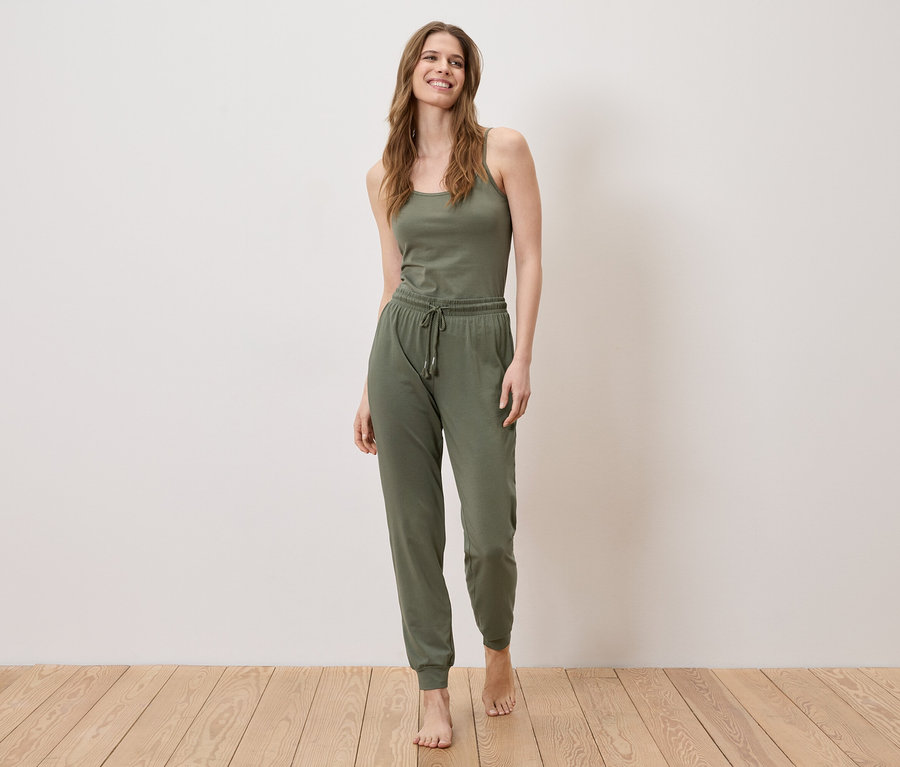 Une femme se tient pieds nus sur un plancher en bois, portant un débardeur et un pantalon de jogging vert olive.