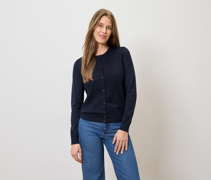 Une femme aux longs cheveux bruns porte un cardigan bleu marine boutonné et un jean bleu sur un fond clair.