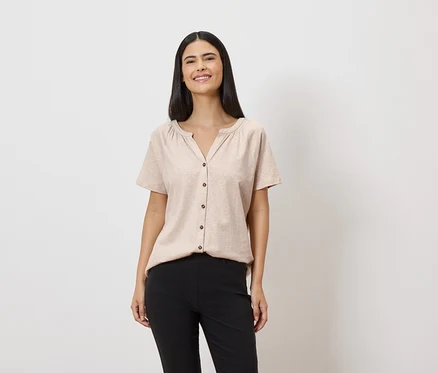 Une femme souriante aux longs cheveux foncés porte un chemisier beige boutonné et un pantalon noir sur fond blanc.