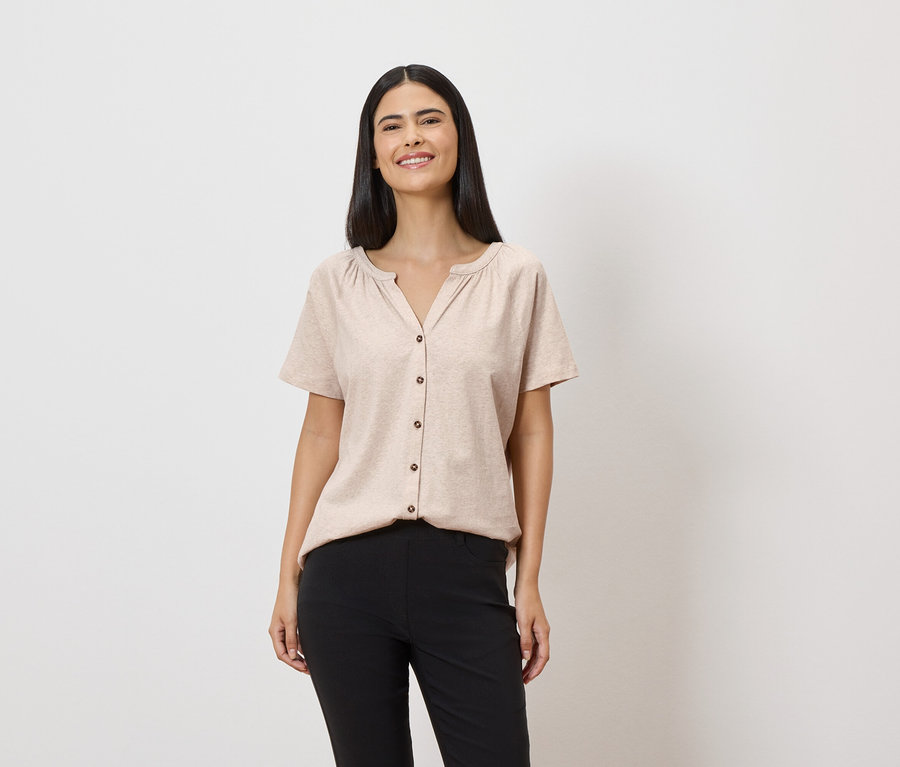 Une femme souriante aux longs cheveux foncés porte un chemisier beige boutonné et un pantalon noir sur fond blanc.