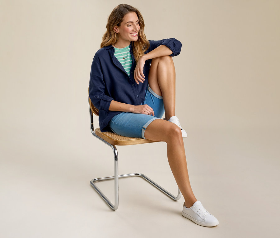 Femme assise sur une chaise portant un t-shirt basique menthe-blanc à rayures sous une chemise bleu marine, un short en jean et des baskets blanches.