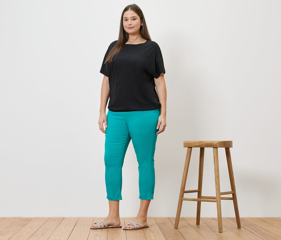 Femme portant un blouse noire et un pantalon stretch 7/8 bleu azur à côté d'un tabouret.