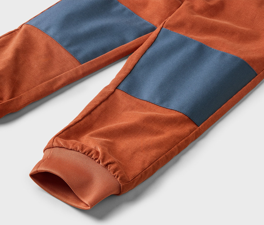 Gros plan d'un pantalon en velours côtelé orange avec des pièces aux genoux bleues et des chevilles élastiquées.