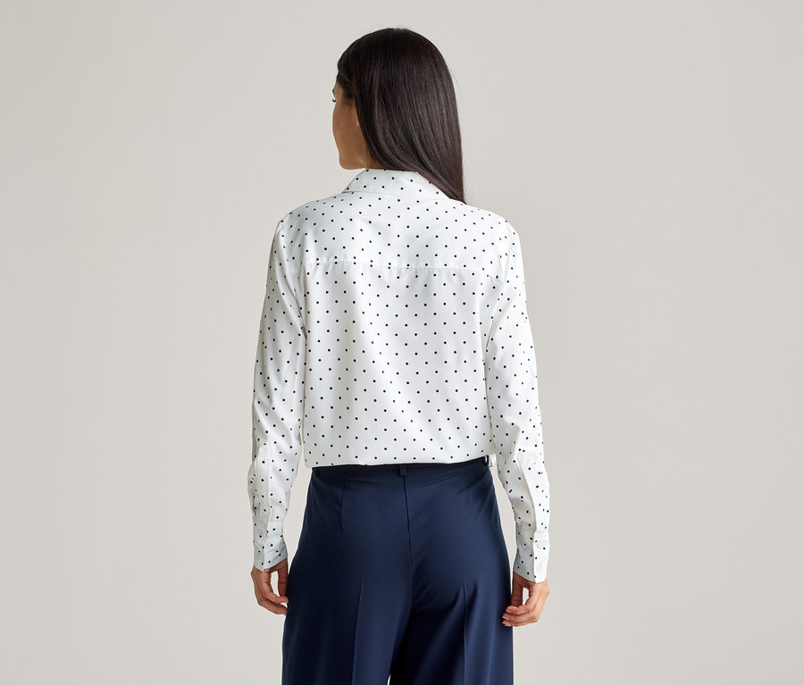 Vue arrière d'une femme aux longs cheveux foncés portant un chemisier blanc à manches longues à pois noirs et un pantalon large bleu marine.