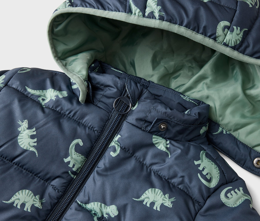 Gros plan d'une veste matelassée bleu marine à capuche avec un motif de dinosaures. L'intérieur de la capuche est vert clair.