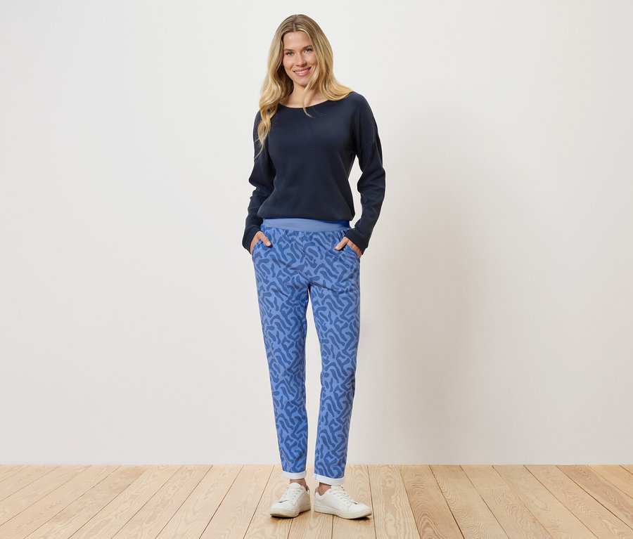Une femme blonde se tient devant un mur blanc, elle porte un pull bleu marine et un pantalon bleu à motifs.