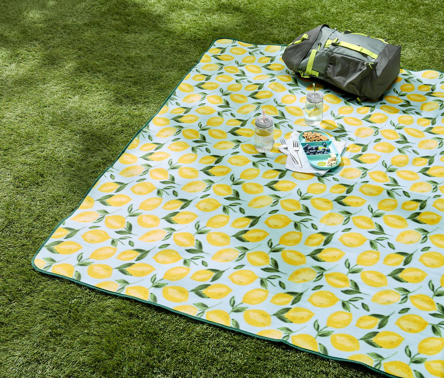 Une couverture de pique-nique à motifs de citron est posée sur de l'herbe verte, avec des boissons, des collations et un sac gris dessus.