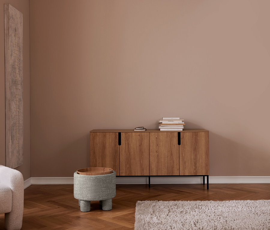 Salon moderne avec murs beiges et parquet à chevrons. Buffet en bois avec livres, tabouret rembourré gris avec plateau en bois, fauteuil gris, tapis clair.