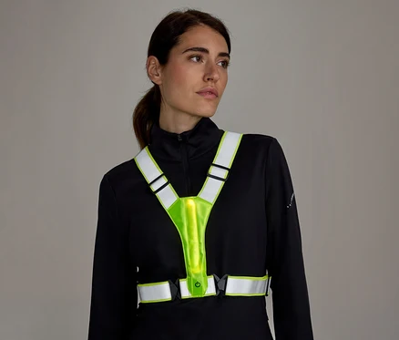 Femme aux cheveux attachés, regardant vers la droite. Elle porte un haut noir à manches longues et un gilet réfléchissant lumineux jaune fluo avec des bandes réfléchissantes blanches.