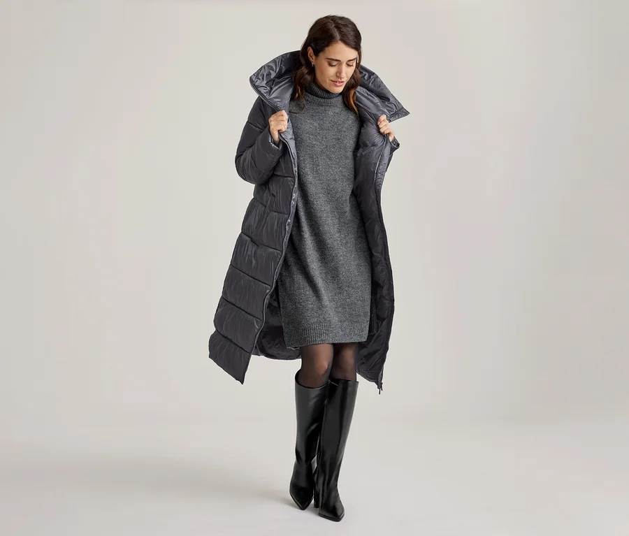 Femme portant un long manteau matelassé gris sur une robe pull grise, des collants noirs et des bottes hautes noires, tenant les revers du manteau.