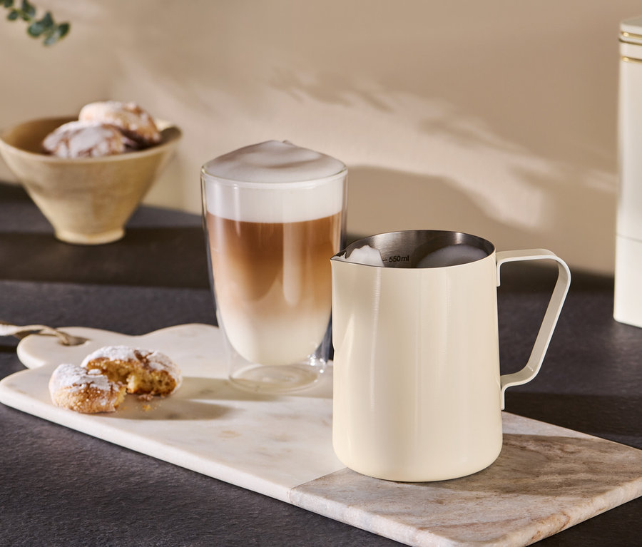 Un pichet à lait beige est posé à côté d'un verre de café sur un plateau en marbre.