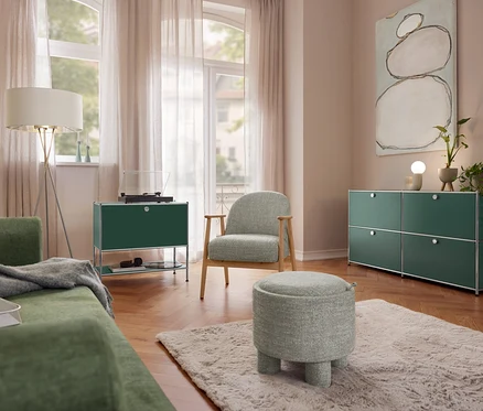 Salon avec canapé vert, lampadaire Eglo « Fondachelli », buffet en métal « CN3 » à 1 abattant et platine vinyle. Un fauteuil avec structure en bois et un pouf sont posés sur un tapis. À droite, un buffet en métal « CN3 » à 4 abattants et une lampe de table en céramique Trio.