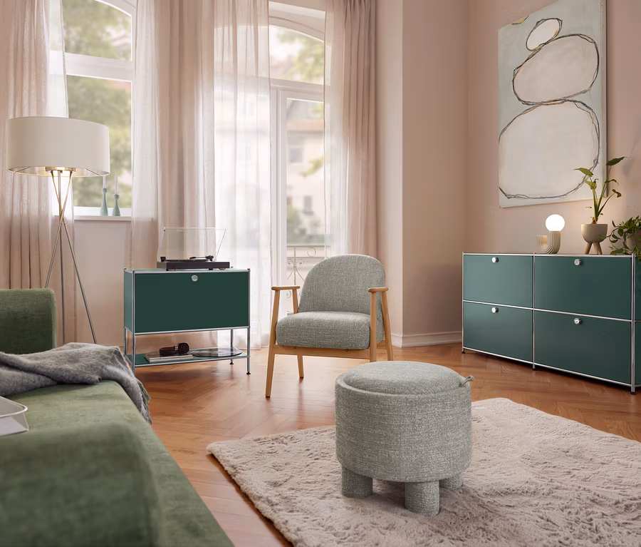 Salon avec canapé vert, lampadaire Eglo « Fondachelli », buffet en métal « CN3 » à 1 abattant et platine vinyle. Un fauteuil avec structure en bois et un pouf sont posés sur un tapis. À droite, un buffet en métal « CN3 » à 4 abattants et une lampe de table en céramique Trio.