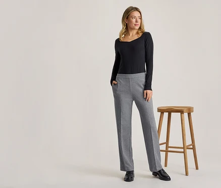 Une femme blonde se tient debout, vêtue d'un haut noir à manches longues et d'un pantalon gris, une main dans la poche, à côté d'un tabouret en bois.
