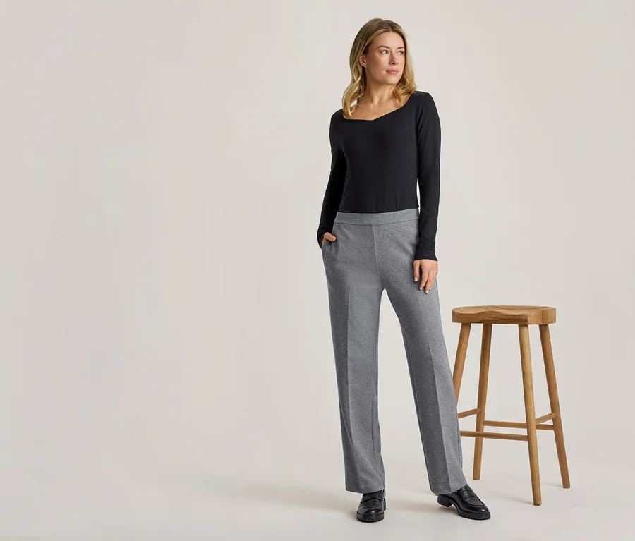 Une femme blonde se tient debout, vêtue d'un haut noir à manches longues et d'un pantalon gris, une main dans la poche, à côté d'un tabouret en bois.