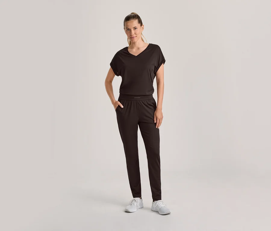 Femme debout, vêtue d'un haut à manches courtes et col en V marron foncé et d'un pantalon assorti, avec des baskets blanches. Sa main gauche est dans sa poche.