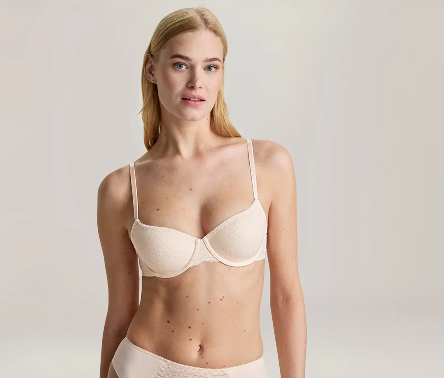 Une femme blonde aux yeux bleus porte un soutien-gorge beige clair et une culotte assortie avec un motif en dentelle, regardant la caméra.