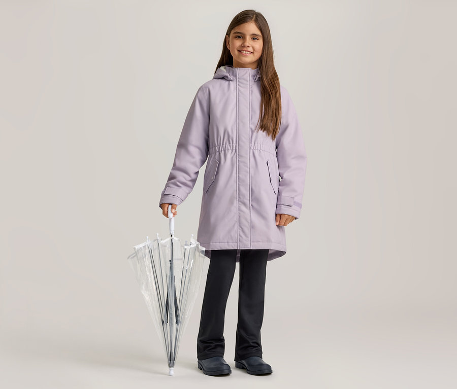 Une fille souriante vêtue d'un manteau lilas, d'un pantalon foncé et de chaussures foncées tient un parapluie transparent fermé.