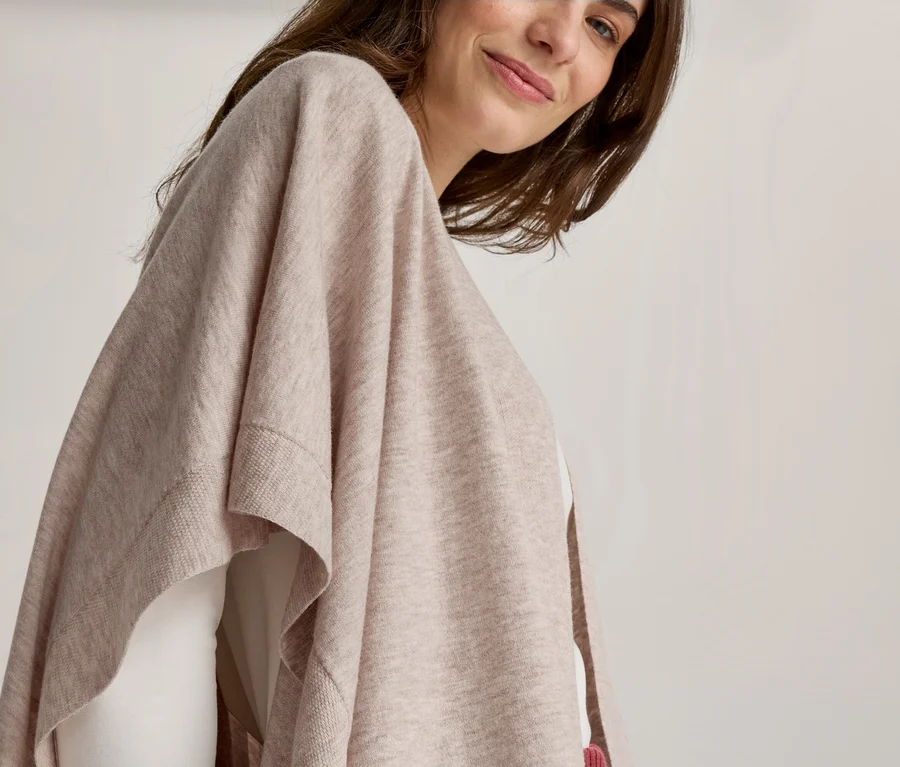 Femme aux cheveux bruns souriante, portant un poncho tricoté beige clair sur un haut blanc.