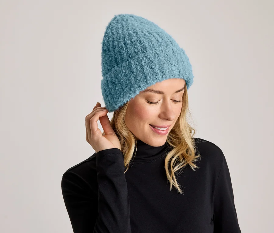 Une femme souriante aux cheveux blonds porte un bonnet tricoté bleu et un col roulé noir. Elle regarde vers le bas et tient une main près de son oreille.
